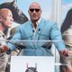 Dwayne Johnson speaking at a lectern in front of two posters for the film "Jumanji"